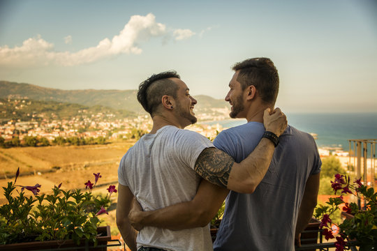 Back view of homosexual couple embracing nd looking at each other on background of resort. 