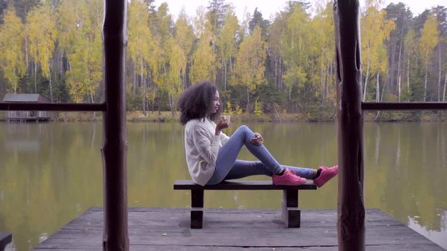 Beautiful girl sitting on the bench and drinking hot tea at the lake