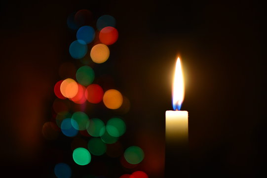Una candela in primo piano che brilla al buio, accompagnata dalle luci soffuse dell'albero di natale sullo sfondo