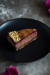 Piece of dark chocolate mousse cake with rose petal and blackberry jam on a black plate and stone background. Vegan, sugar, gluten free, dairy dessert. Vertical