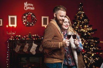 Attractive happy couple on Christmas day standing in front of the decorated tree smiling lovingly into each others eyes and holding glasses with red wine