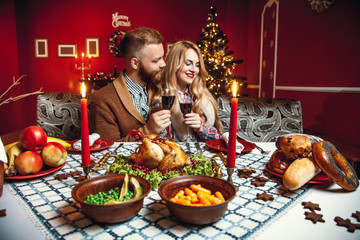 Beautiful couple in a decorated festive interior with a Christmas tree drinking wine. A romantic dinner for thanksgiving with fried chicken and candles