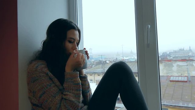 Close up view of a young beautiful girl sitting on a windowsill at the window overlooking an old european city and drinking hot coffee or tea from a mug.