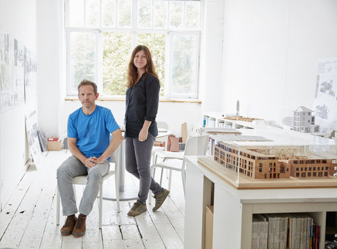 A Modern Office With White Walls And Floors. A Man Sitting And A Woman Standing. An Architectural Model On A Table. 