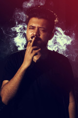 Young man smoking cigarette on dark background looking into came
