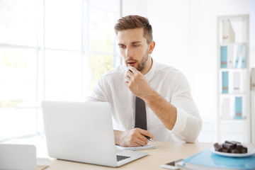 Young man eating candies while working with laptop in office