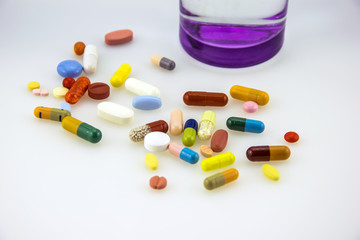 A lot og colored tablets and capsules near a glass of water with purple bottom isolated on white background.