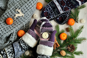 Christmas time, clock in knitted gloves hands. Tangerine and fir branches close up over rustic wooden background