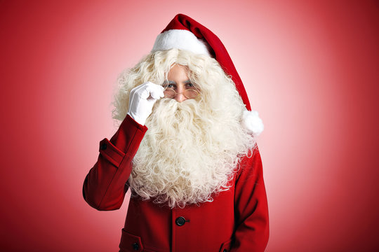 Santa Claus with a kind smile and a large white beard in red coat and hat and white gloves adjusting his glasses on red background
