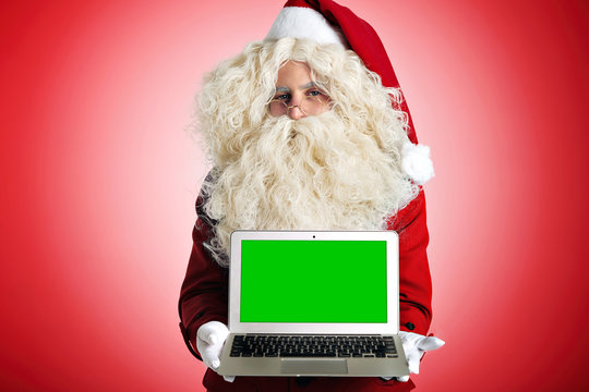 Santa looks at camera and offers opened new portable laptop with your own picture on screen instead chromakey color, isolated on red. Computer repairing or programming holiday concept