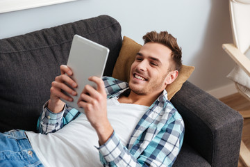 Cheerful bristle man lies on sofa and using tablet computer
