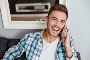 Young laughing man sitting on sofa and talking by phone