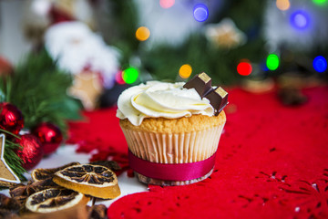 Пирожное, стоит на красная новогодней салфетке на белом деревянном столе в окружении зеленой новогодней гирлянды и огней, игрушки Санта клаус, имбирных пряников в виде звезды, гранат, сушеного лимон - obrazy, fototapety, plakaty