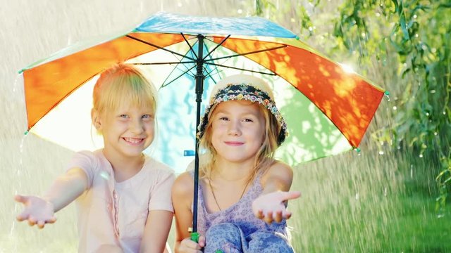 Two carefree girl sit under an umbrella, hiding from the warm summer rain. The concept - a happy childhood
