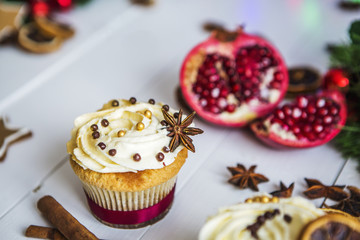 Пирожное с кремом, капкейк, мбирный пряник в виде звезды, разрезанный красный гранат, корица, сушеные лимоны лежат на белом деревянном столе на фоне зеленой новогодней гирлянды и новогодних огней.