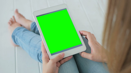 Woman sitting on the white wooden floor and using vertical tablet computer with green screen. Close up shot of woman's hands with pad. Technology and internet concept