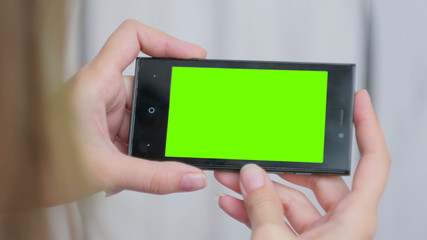 Woman looking at horizontal smartphone with green screen. Close up shot of woman's hands with mobile. White wooden background