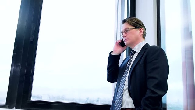 Businessman talks on the phone standing by a large window. Slow motion.
