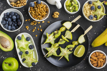 Healthy fresh breakfast. Granola, quinoa bowl, various nuts and fruit skewers with green fruits on a dark background. Top view. Color year. Greenery
