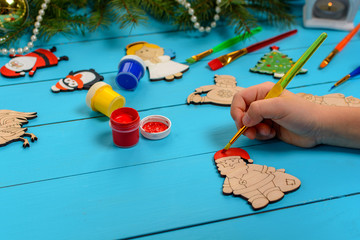 Boy decorates by paints handmade wooden Christmas decorations on blue vintage table with a Christmas tree and toys