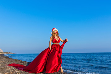 Beautiful woman in red dress and Santa hat is on the shore of the sea