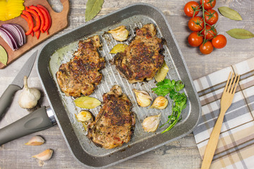 Fried meat slices in the pan, tomatoes greens onions garlic pepper, tasty food. Knife fork, on wooden table, background.