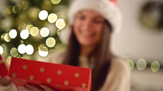 Pretty Woman in Santa Hat Astonished with the Surprise Inside of the Red Box. Cute Smiling Girl opening Christmas Gift on Xmas Tree Background, Closeup.