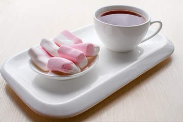 Fototapeta na wymiar On the table a Cup of tea and pink and white marshmallows in a white ceramic dish. Breakfast on a light background. High key. Selective focus. For culinary publishers and websites.