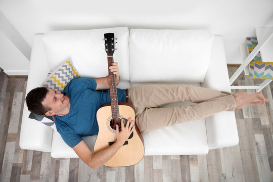 Young man playing guitar and lying on the sofa in light room