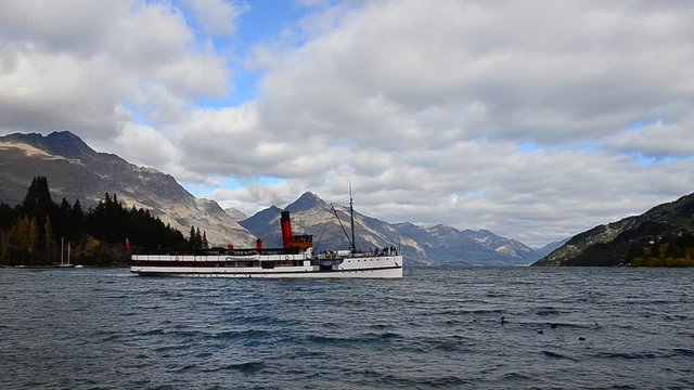 The iconic TSS Earnslaw steamship sails across Lake Wakatipu in Queenstown, New Zealand, surrounded by the majestic Southern Alps under a dramatic cloudy sky.