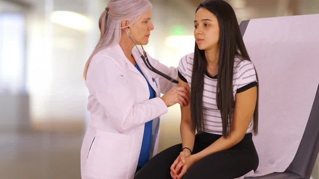 A young girl receives her check-up from a medial professional. A teen girl gets her check up from an elderly doctor. 