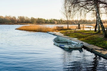 Small water filled rowboat moored beside a narrow wooden pier on a cold winter morning. Backlit scene with reed and coastline in background. Ice inside the boat. Copy space.