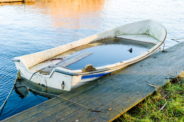Small water filled rowboat moored beside a narrow wooden pier on a cold winter morning. Ice inside the boat.