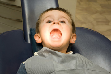 Funny five y.o. boy with wide opened mouth is sitting in the dentist chair under the medical lamp light ready to treat his teeth - 130366398