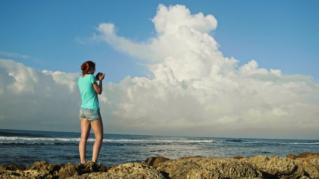 Young woman with red hair in glasses photographs sea standing on the rocks , beach of Dominican Republic