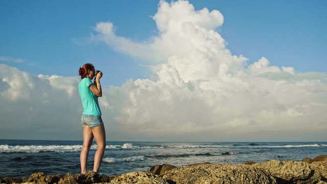 Young woman with red hair in glasses photographs sea standing on the rocks , beach of Dominican Republic, wide angle
