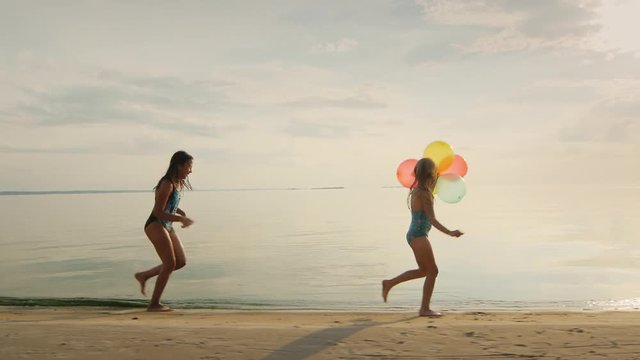 Two sisters playing together on the beach. One girl runs after another, holding balloons