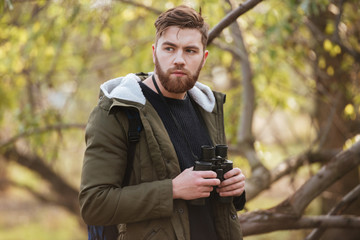 Serious bearded man holding field-glass standing in the forest