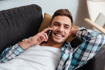 Laughing bristle man lies on sofa while talking by phone