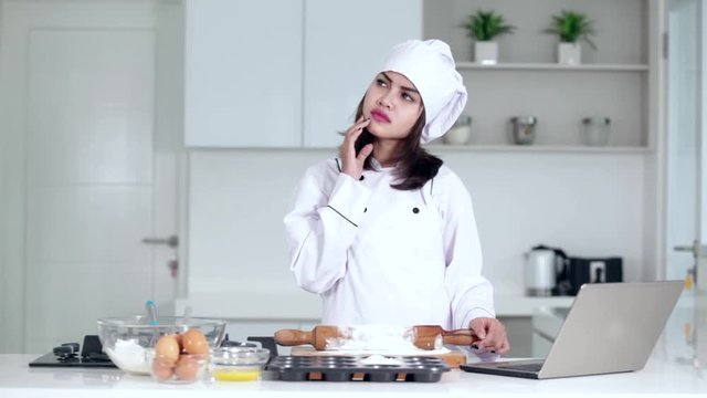 
Young woman making cake in the kitchen while thinking idea and browsing internet on the laptop
