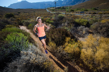 Wide angle view of a fit trail running female athlete running on a trail in the hills with dry shrubs on a bright sunny day