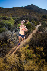 Naklejka premium Szeroki kąt widzenia sprawnej zawodniczki biegnącej w terenie biegającym po szlaku na wzgórzach z suchymi krzewami w jasny, słoneczny dzień