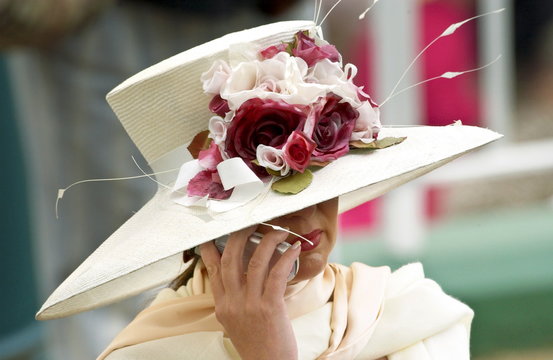 Race-goer Wearing A Cream Hat Trimmed With Flowers At Royal Ascot Races While Chatting On Her Mobile Cell Phone.