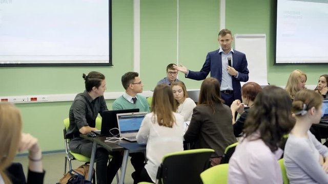 Group of students in classroom at the university are listening attentively the lecture on Economics of famous lecturer. Speaker in blue suit with the microphone is very positive and delivering