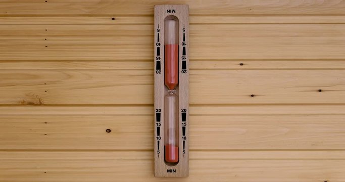 Zeitraffer einer typischen Sanduhr in einer h&ouml;lzernen skandinavischen Sauna / H&ouml;lzerne Sanduhr an einer mit Holz vert&auml;felten Wand mit orangenfarbenem Sand und schwarzer Minutenskala.