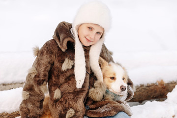 Happy little girl in a white fur coat and hat sitting on logs in the winter and embraces a red and white dog