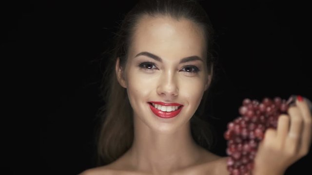 Beautiful young brunette woman with red lipstick holding grapes and looking at camera