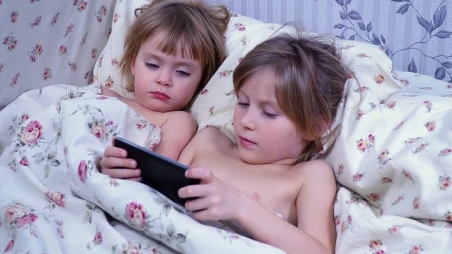 Two beautiful children lying in bed under the blanket and look at phone, closeup