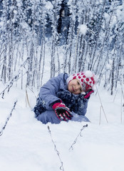 Fototapeta na wymiar Маленькая девочка гуляет в зимнем лесу. Ребенок сидит в снегу и улыбается.