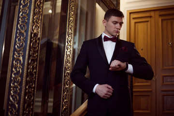 Handsome young man in a tuxedo looking at the camera. Interior of the hotel. Clothing for the festive evening. He looks at his watch